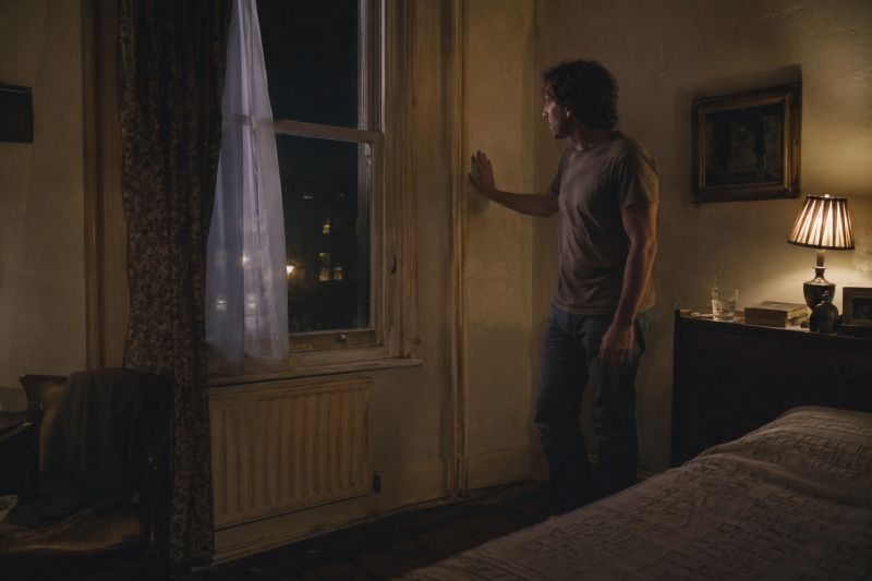 A man standing in a dim bedroom with one hand against the wall beside a window and radiator.